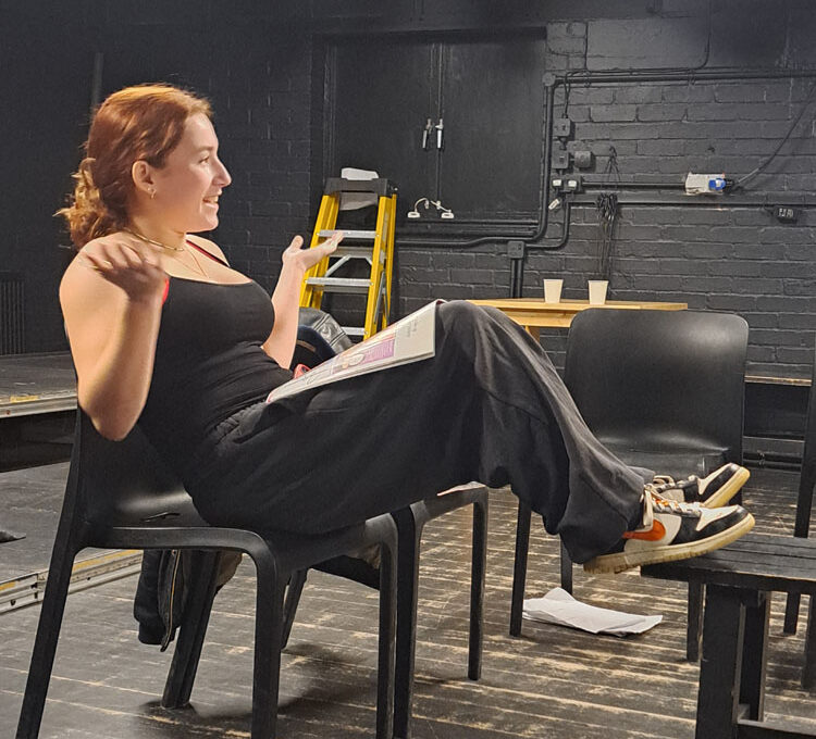 A girl dressed in black top and trousers sits on a chair on a stage reading a magazine