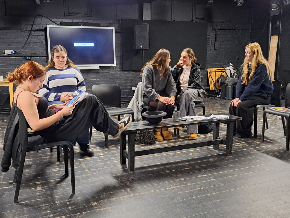 Five girls sit on chair around a low table as they perform a play in the rehearsal space at The King Alfred School North London