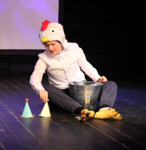 A student dressed in a chicken hat and chicken feet sits on the floor with a bucket and two party hats