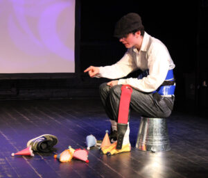 A man sits on a bucket wearing a bird-foot shaped shoe pointing at a shoe and party hats on the floor in front of him