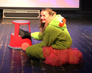 A student on stage wearing a pink tutu and green onesie grins at the audience
