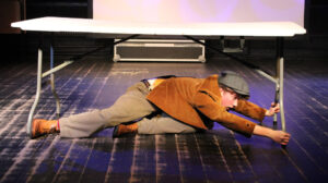 A student performs on stage at The King Alfred School North London dressed in a tweed jacket and flat cap they try to prop up the legs of a table