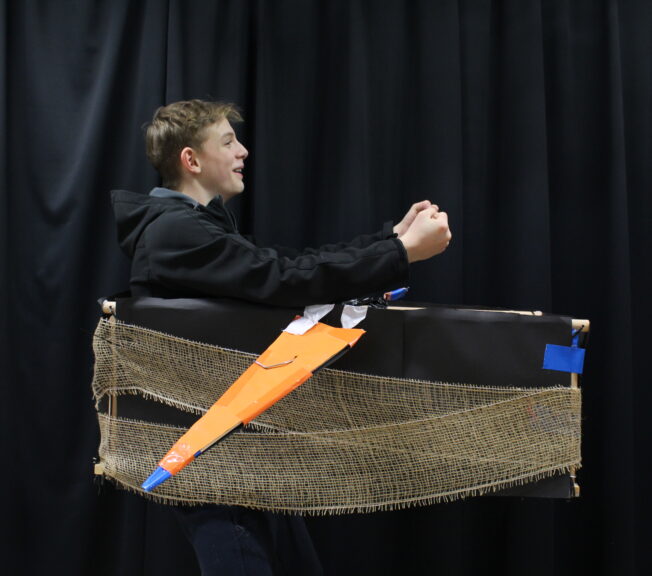 A student wearing a box shaped like a plane around his waist, with orange wings and a black body