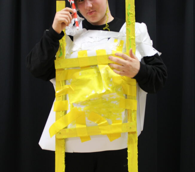 A male student wears a white tabard with strips of yellow paper across the front, giving the illusion of a fried egg