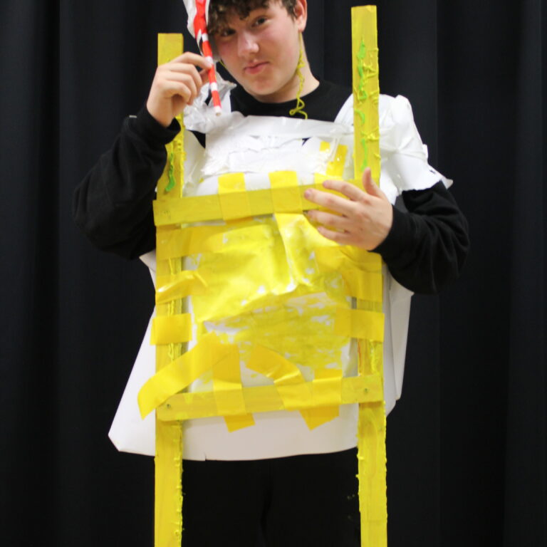 A male student wears a white tabard with strips of yellow paper across the front, giving the illusion of a fried egg