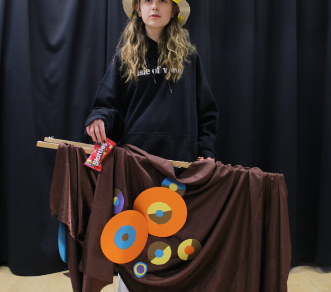 A student wears a paper top hat decorated with abstract circles, and a wooden frame around her waist draped with brown fabric and matching circles