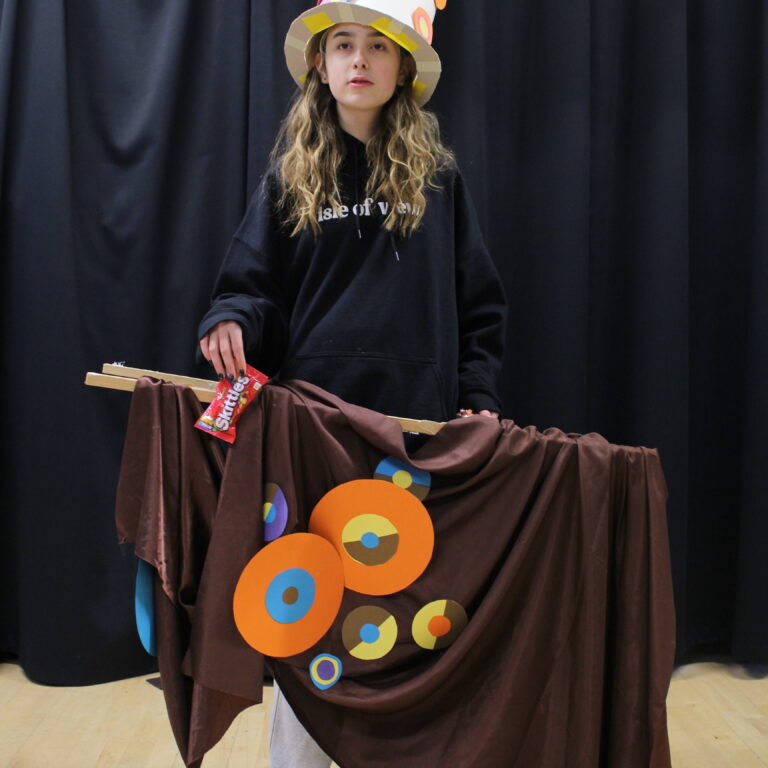 A student wears a paper top hat decorated with abstract circles, and a wooden frame around her waist draped with brown fabric and matching circles