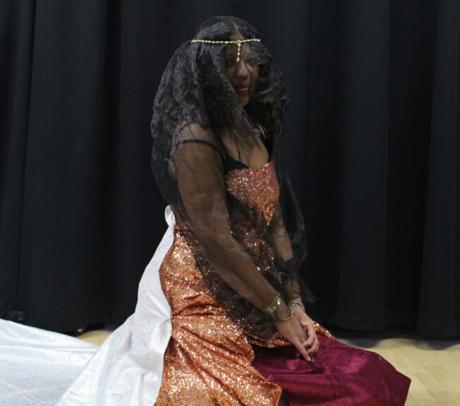 A student kneels on the floor, wearing a self-made outfit featuring a black veil, salmon pink sequin dress and white train