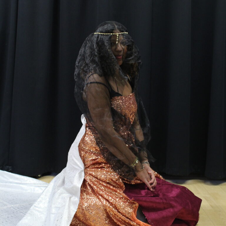 A student kneels on the floor, wearing a self-made outfit featuring a black veil, salmon pink sequin dress and white train