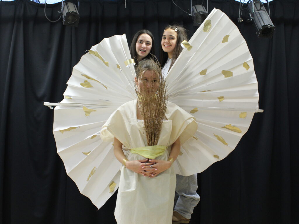 A student wears a homemade white goddess-style dress with rounded white wings with gold sports. She holds a bunch of sticks over her face, and two students stand behind her.