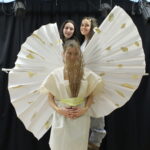 A student wears a homemade white goddess-style dress with rounded white wings with gold sports. She holds a bunch of sticks over her face, and two students stand behind her.