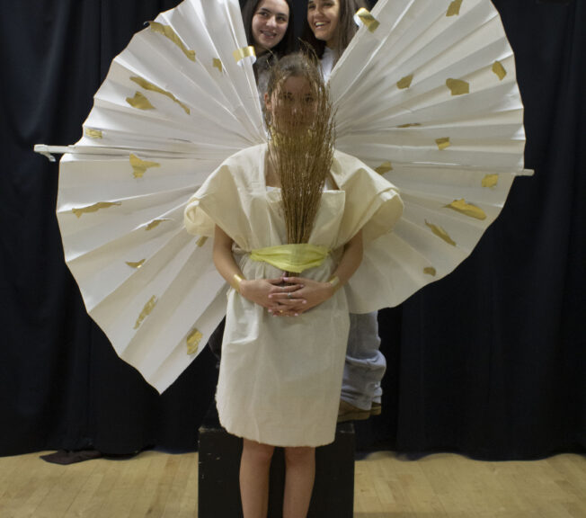 A student wears a homemade white goddess-style dress with rounded white wings with gold sports. She holds a bunch of sticks over her face, and two students stand behind her.