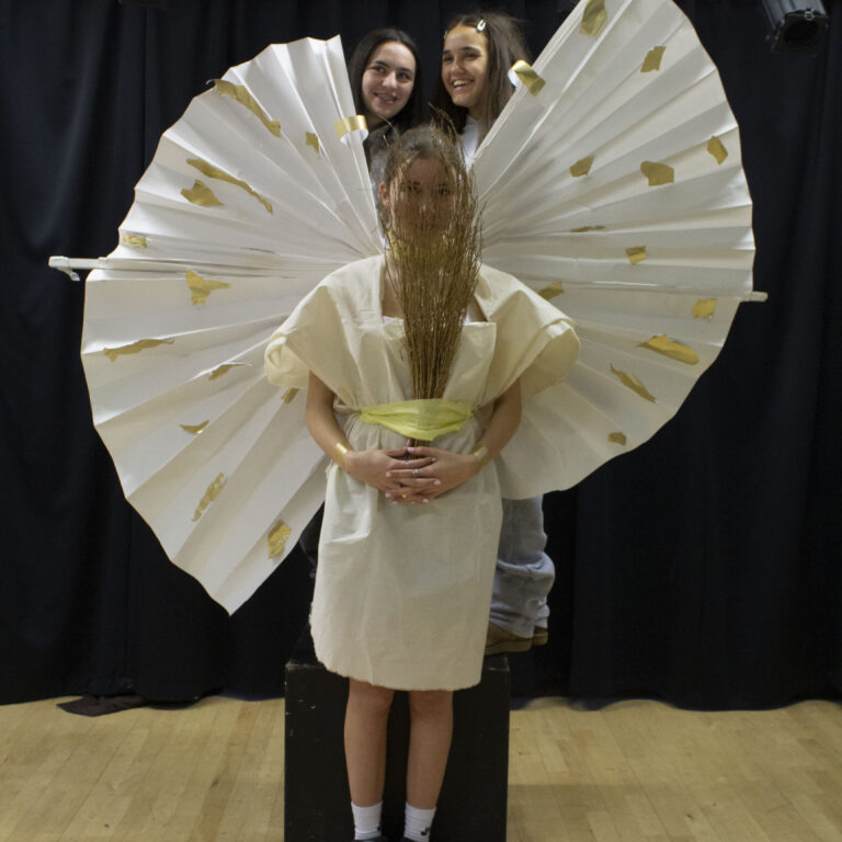 A student wears a homemade white goddess-style dress with rounded white wings with gold sports. She holds a bunch of sticks over her face, and two students stand behind her.