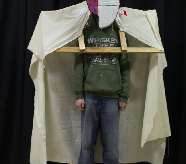 A student wears a design inspired by abstract art. It features a wooden structure over his shoulders draped with white material, and a white and pink abstract headpiece.