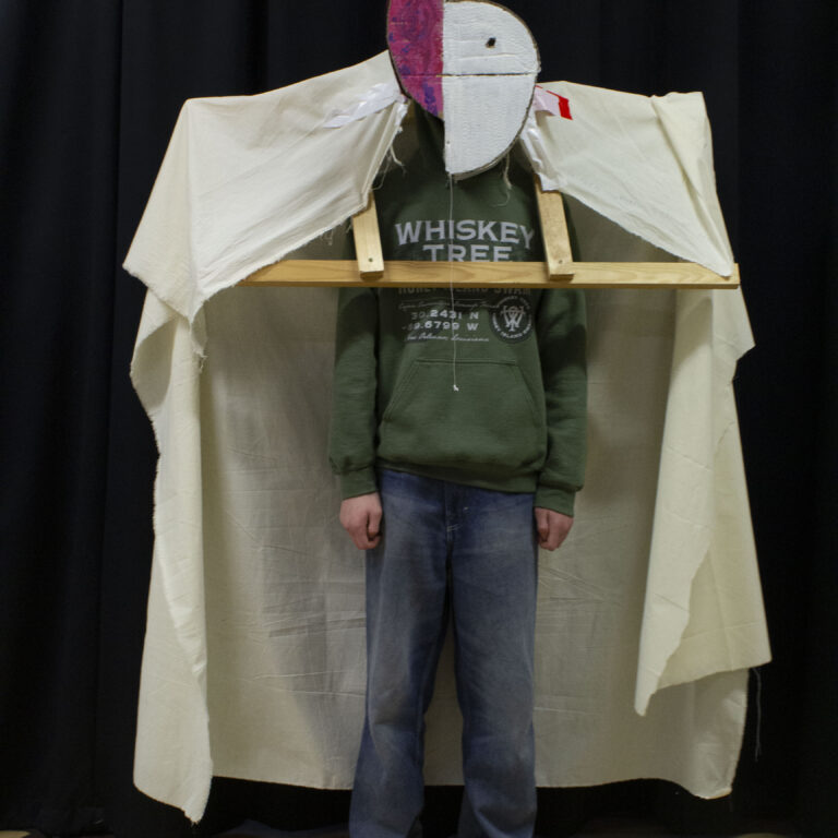 A student wears a design inspired by abstract art. It features a wooden structure over his shoulders draped with white material, and a white and pink abstract headpiece.