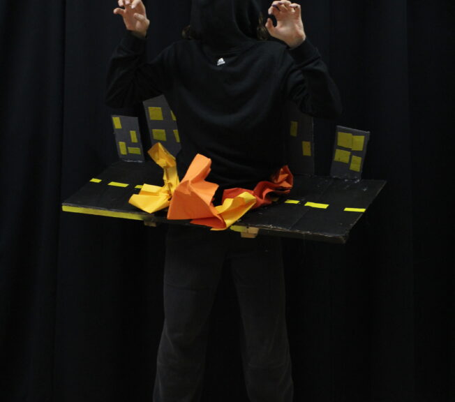A student dressed all in black wears an abstract design around his waist, depicting a city scene with flashes of flames on the road.