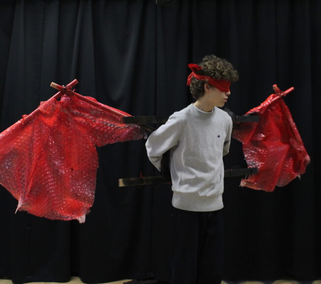 A student wears a large pair of red wings, made from wood and bubble wrap spraypainted red. The student also wears a red blindfold.