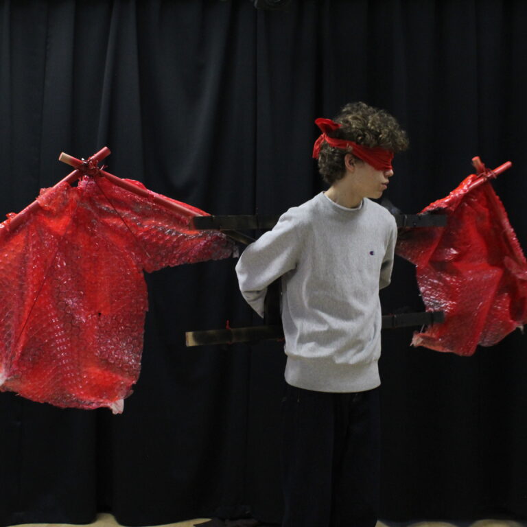 A student wears a large pair of red wings, made from wood and bubble wrap spraypainted red. The student also wears a red blindfold.