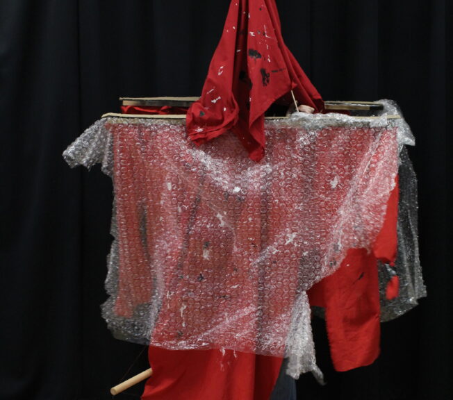 A student wears a wooden structure draped with red fabric and bubble wrap, topped with a matching red fabric hat