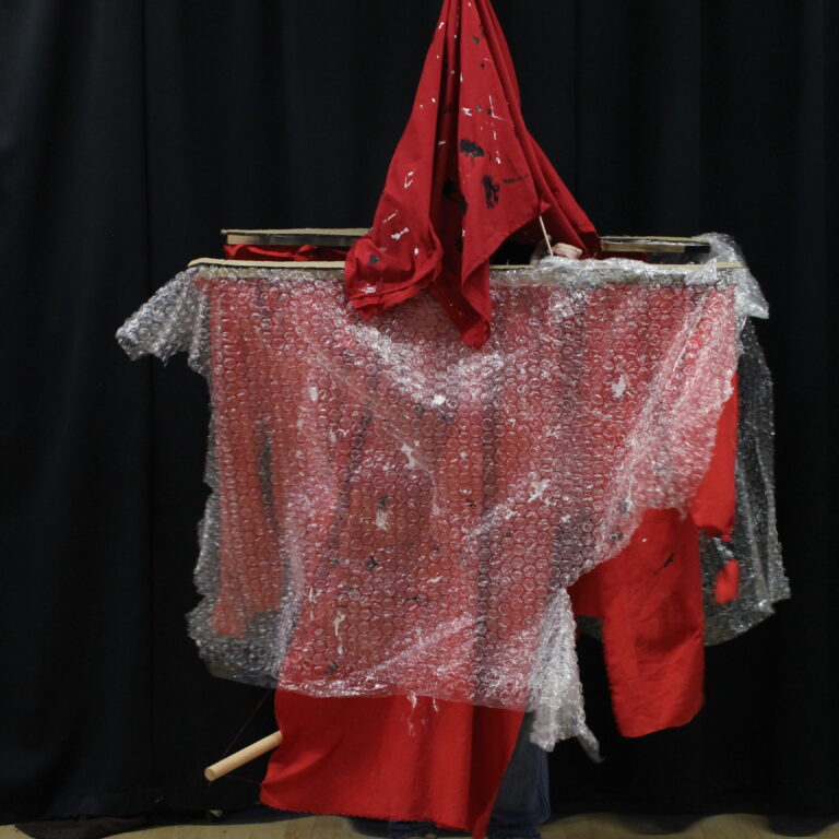 A student wears a wooden structure draped with red fabric and bubble wrap, topped with a matching red fabric hat