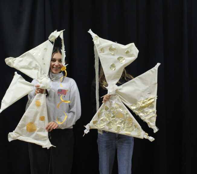 Two female students stand side by side, each holding an abstract structure featuring white sails with gold applique