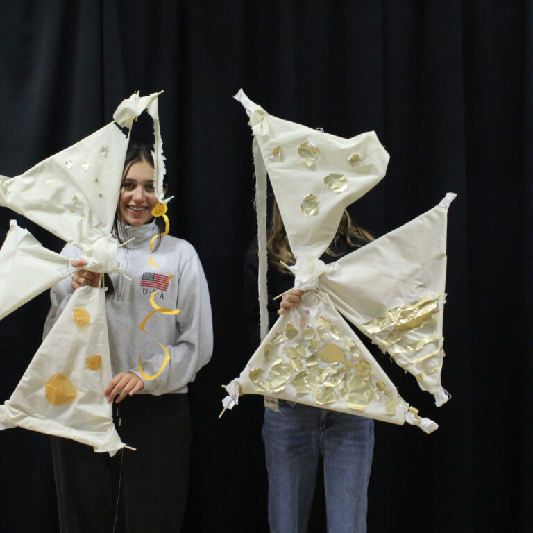 Two female students stand side by side, each holding an abstract structure featuring white sails with gold applique