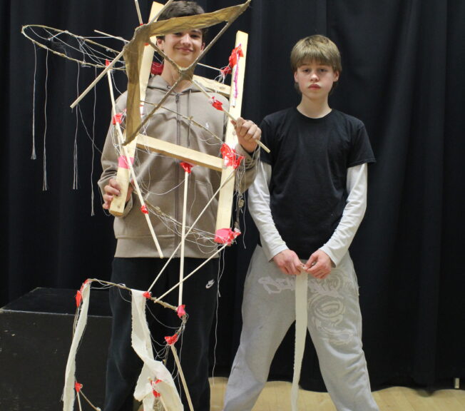 Two male students stand side by side. One is wearing a wooden structure with wires and string attached to it.