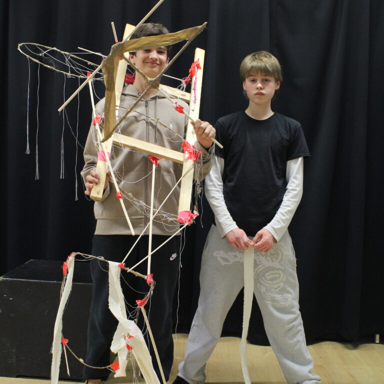 Two male students stand side by side. One is wearing a wooden structure with wires and string attached to it.