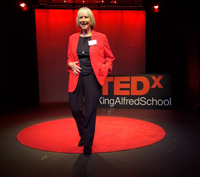 Woman wearing a black top and trousers with a red jacket. She is standing on a black stage with a round red carpet and a sign reading 'TEDx King Alfred School'