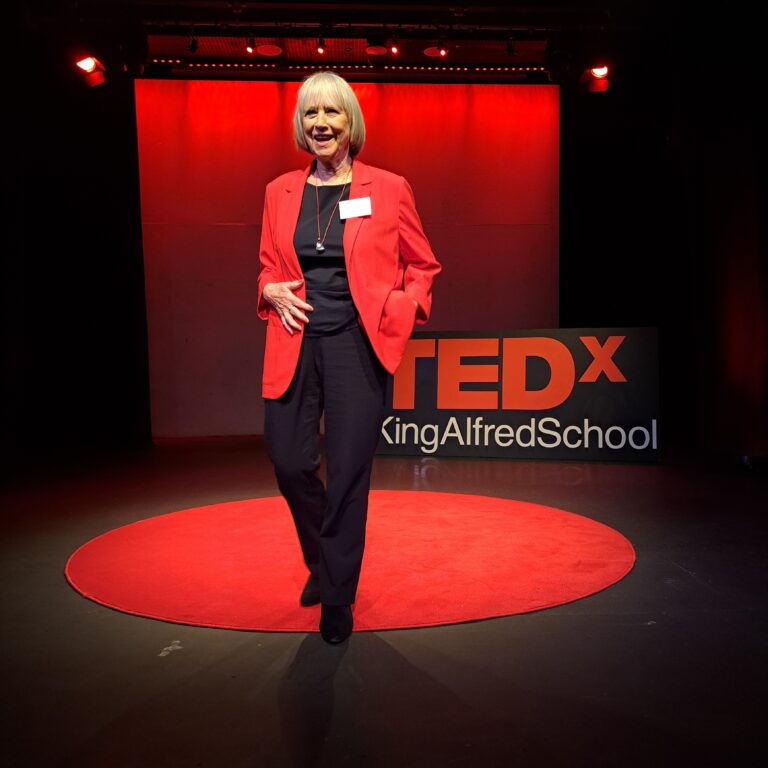 Woman wearing a black top and trousers with a red jacket. She is standing on a black stage with a round red carpet and a sign reading 'TEDx King Alfred School'