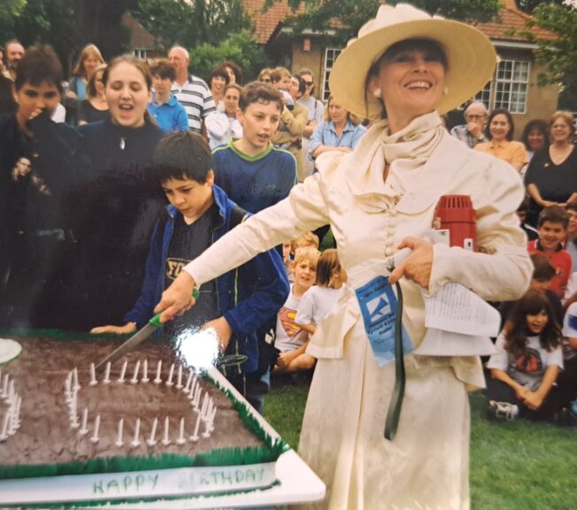 Archive photograph showing a woman dressed as Millicent Fawcett cutting a cake with candles arranged to spell '100' in front of a crowd of students on the field at The King Alfred School