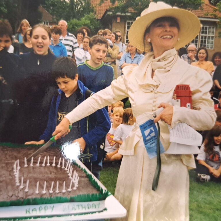 Archive photograph showing a woman dressed as Millicent Fawcett cutting a cake with candles arranged to spell '100' in front of a crowd of students on the field at The King Alfred School