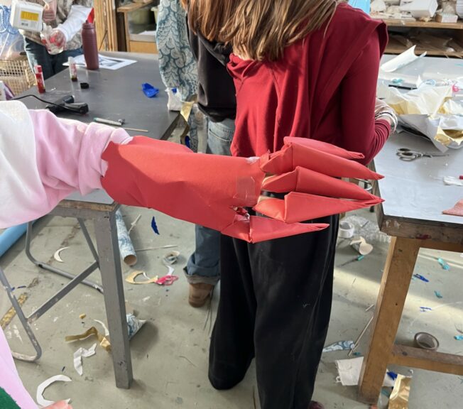 A student models a 'claw' and cuff made from red paper