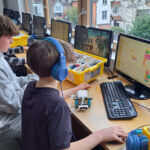 Two students, one wearing blue headphones, sit at a desk, using a computer to use a coding programme