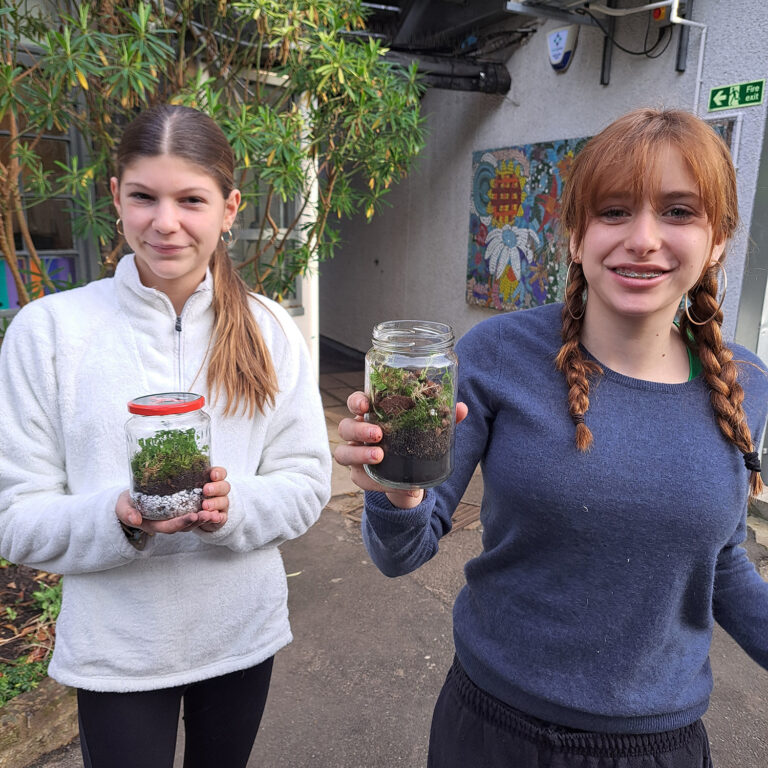Two female students stand outside holding terrarium in glass jars