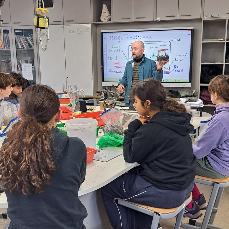 A male teacher holding a terrarium stands in front of students sat at science benches