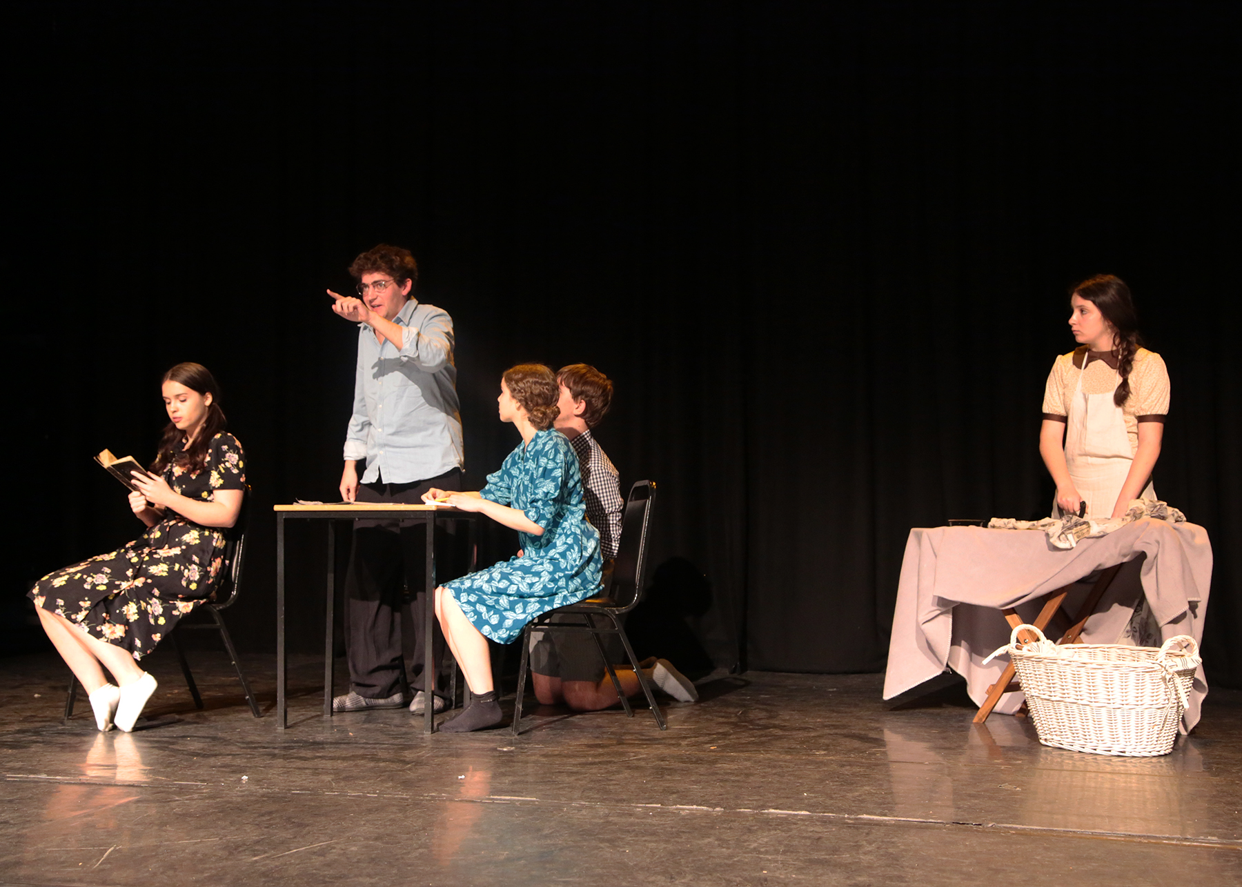 Students on stage during a French language performance. One stands ironing at an old-fashioned ironing board, three sit around a table, and one stands at the table, pointing an accusatory finger in the direction of the audience.