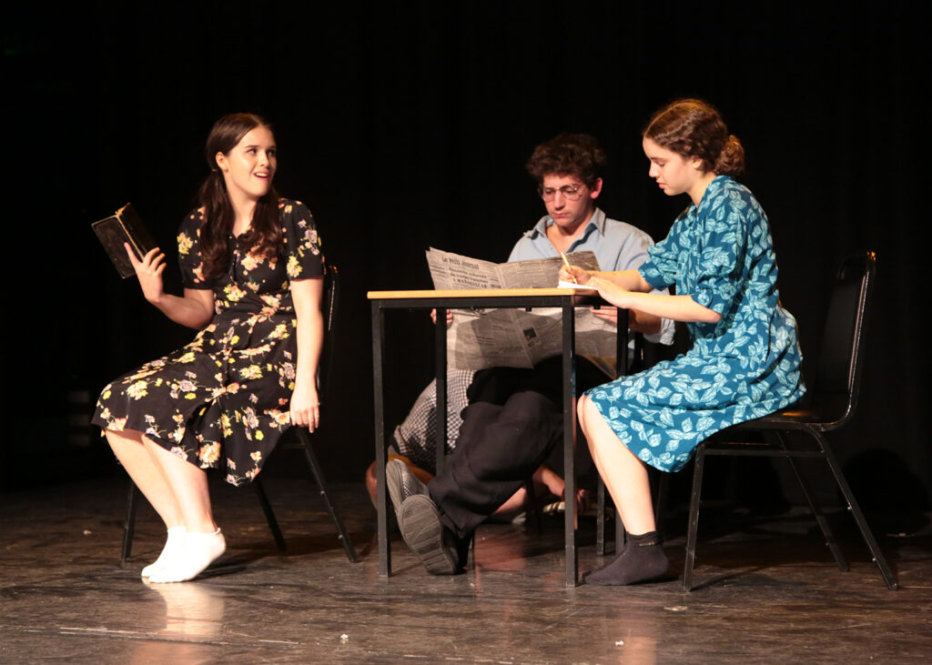 Three students sit around a table on stage. One reads a newspaper, one is writing, and one is reading a book and looking incredulously at the others