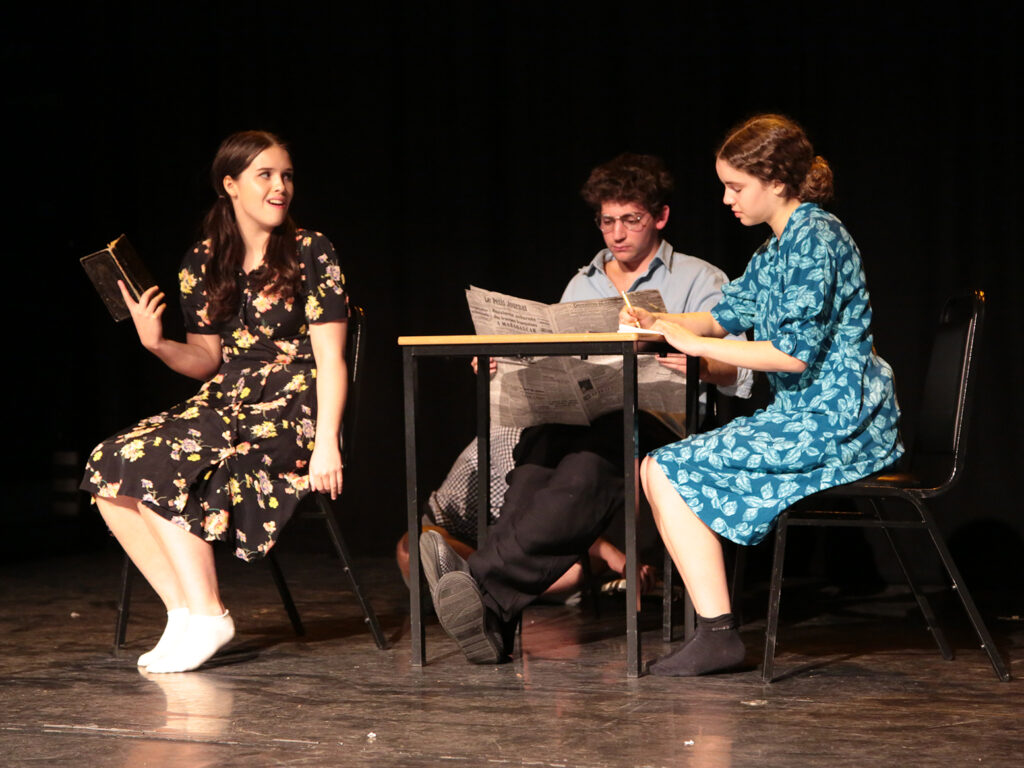 Three students sit around a table on stage. One reads a newspaper, one is writing, and one is reading a book and looking incredulously at the others