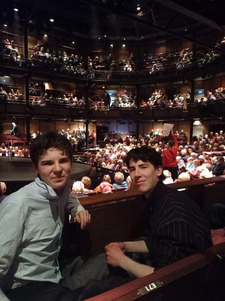 Two male Sixth Form students smile at the camera with a packed theatre behind them
