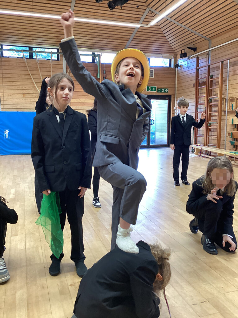 Young students wearing suits rehearse in a school hall. One has their knee raised with their foot on the back of someone crouched-down.