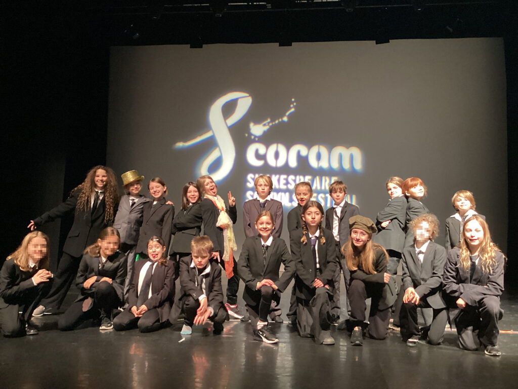 Young students wearing white shirts and black suits stand or kneel on a theatre stage, with a black backdrop reading 'Coram Shakespeare Schools Festival' behind them