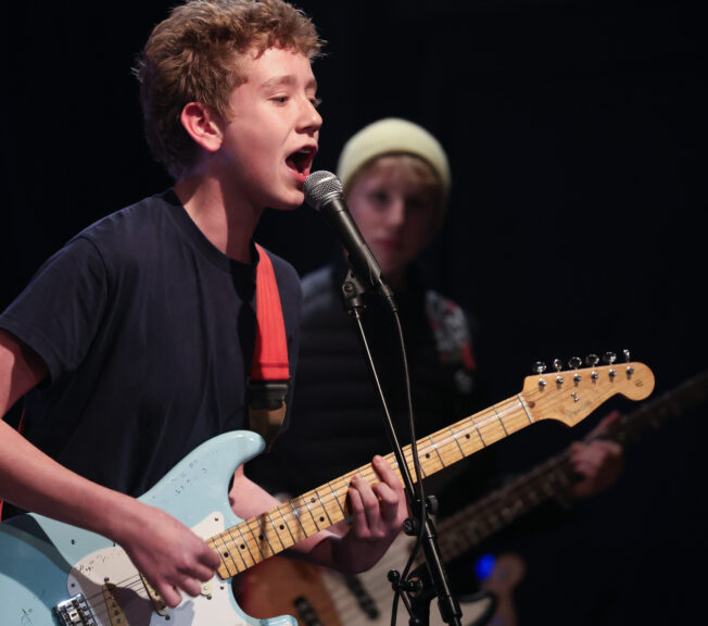 A male student wearing a black t-shirt plays a blue electric guitar whilst singing into a microphone