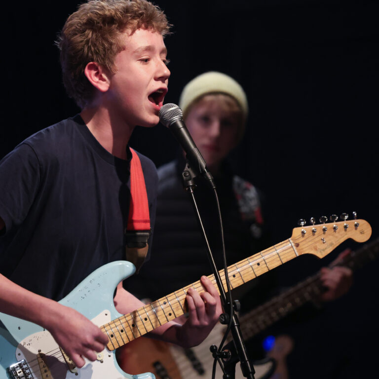 A male student wearing a black t-shirt plays a blue electric guitar whilst singing into a microphone