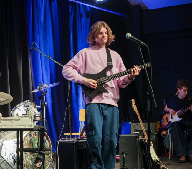 Male students play instruments (one on drums, two on electric guitar) on stage