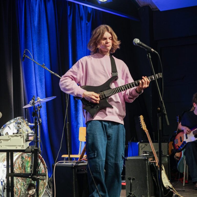 Male students play instruments (one on drums, two on electric guitar) on stage