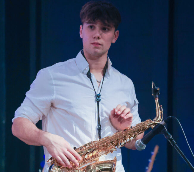A male student wearing a white shirt and black trousers holds a saxophone on stage during a concert
