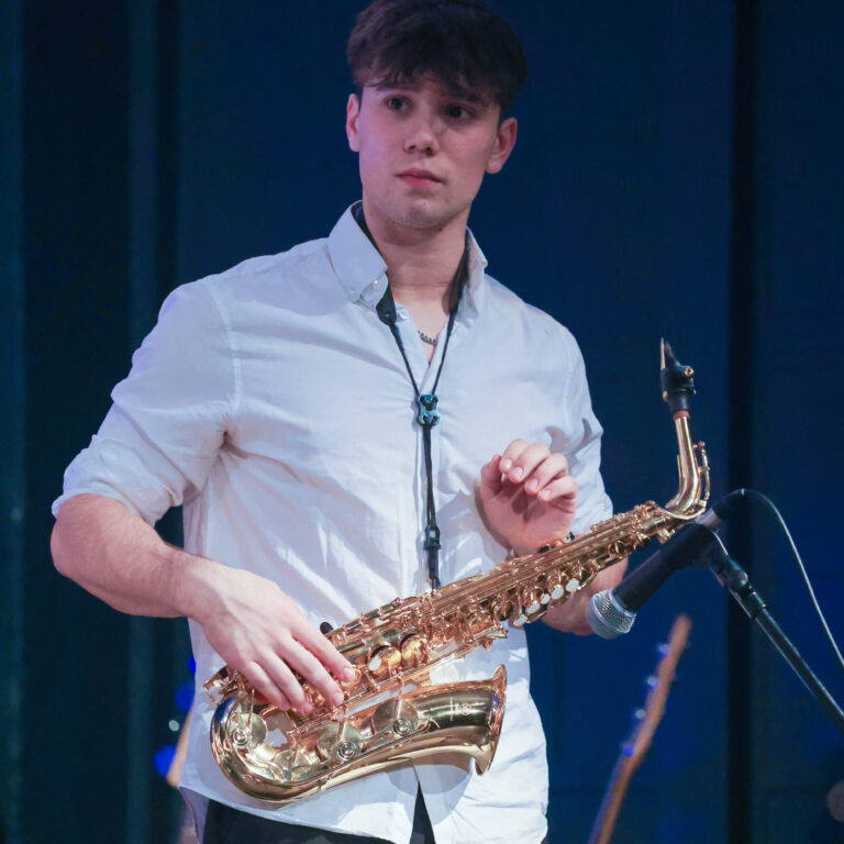 A male student wearing a white shirt and black trousers holds a saxophone on stage during a concert