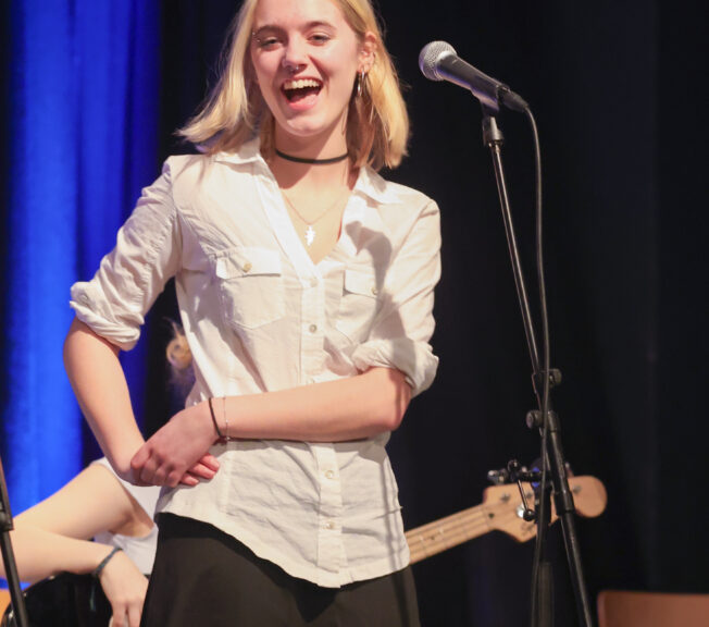 A female student wearing a white shirt and black skirt laughs whilst standing in front of a microphone on stage