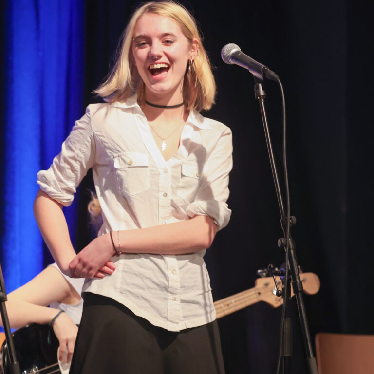 A female student wearing a white shirt and black skirt laughs whilst standing in front of a microphone on stage